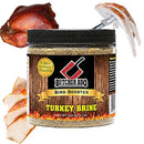 A jar of Butcher BBQ Bird Booster Turkey Brine shown on a white background with a whole roasted turkey and sliced cooked turkey pieces used as contextual food props. The product is a clear plastic jar with a black lid and a wood-grain label featuring the brand name 'Butcher BBQ' and the text 'Bird Booster' and 'Turkey Brine'.