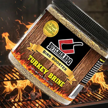 A jar of Butcher BBQ Bird Booster Turkey Brine seasoning is shown at an angle on a grill with flames and coals. The label features a wood-grain texture, a black background with a red logo, and a gold seal that reads '2x Strong Bird Booster'.