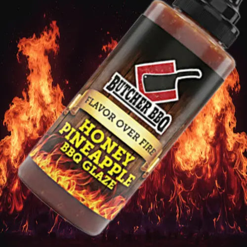 A bottle of Butcher BBQ Honey Pineapple BBQ Glaze shown at an angle against a dramatic background of orange and yellow flames. The bottle has a black cap and a black label with red, white, and yellow text and graphics.